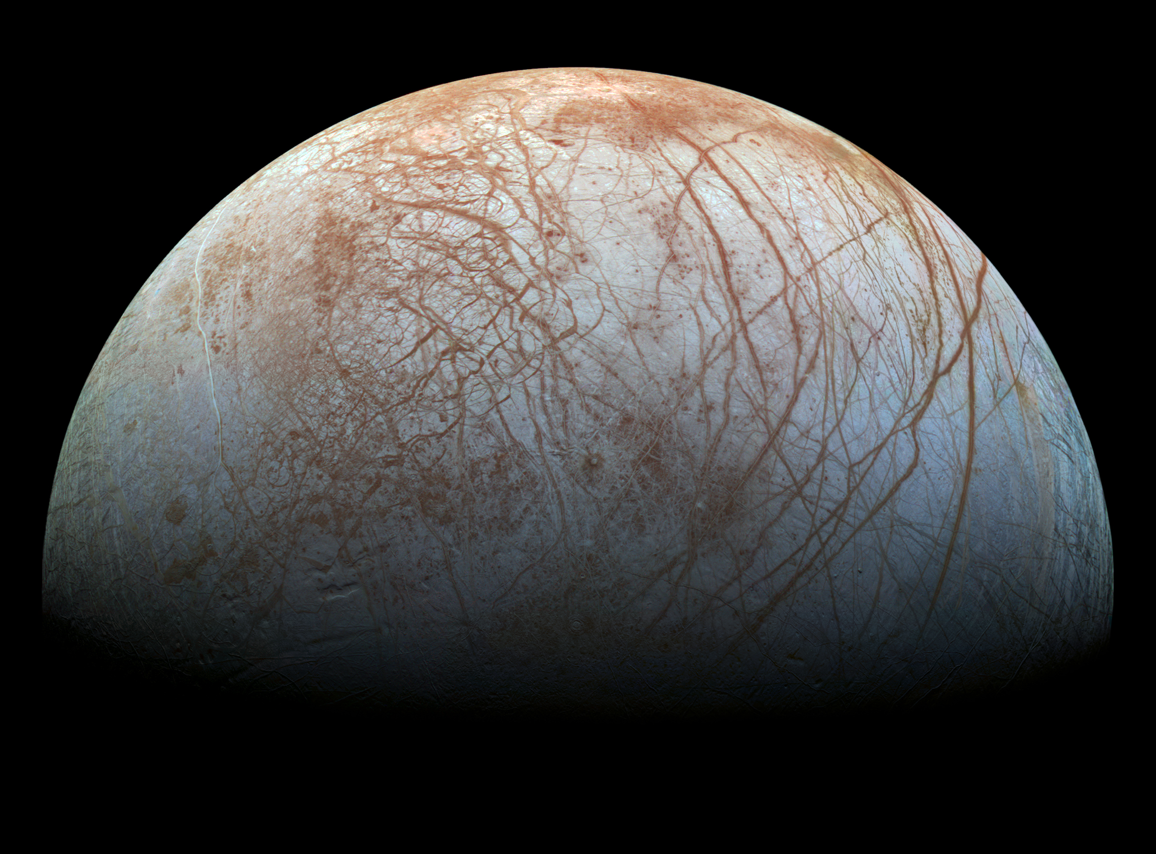Europa is visible as a half-sphere resting upon the dark black background of space. Europa's surface is off-white in color, with a slight blueish hue. Long, dark rusty lines are visible across the surface of the moon. On the left side of the moon, the lines are in a visibly tighter patterns, and on the right side of the moon the lines are deeper and extend across the length of the moon in long curving acs. As more of the moon is visible at the bottom of the image, it fades into darkness. 