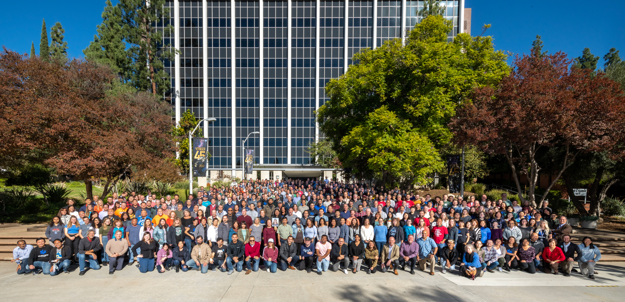 Dozens of people stand on a stair case in front of a glass building, looking towards the camera. They are posing for a team image, with some kneeling in front. They are framed on the top left and right be trees, with a blue sky in the distance. The image was taken at JPL in Pasadena, California. 